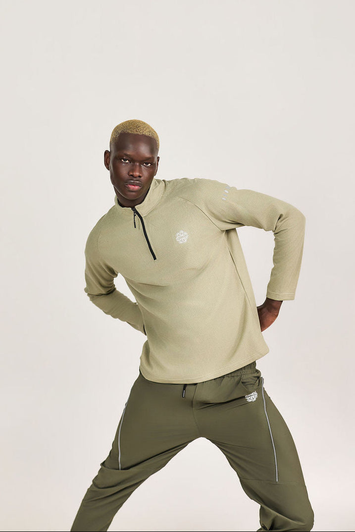 Man wearing a beige long-sleeve shirt and green pants on a light background